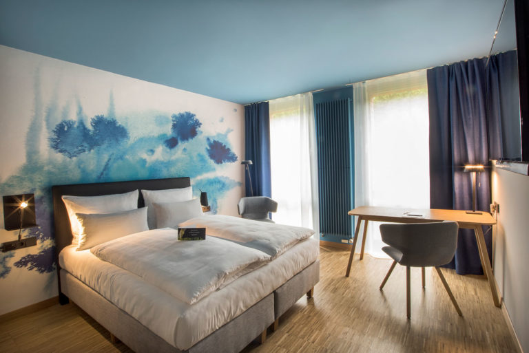 Die Arcadeon Suite bietet ein modernes Hotelzimmer mit einem Doppelbett, abstrakter blauer Wandkunst, Holzboden und einem Schreibtisch mit Stuhl. Elegant drapierte Vorhänge über großen Fenstern schaffen ein einladendes Ambiente.