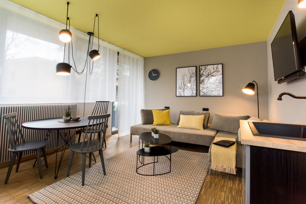 Modernes Wohnzimmer im ARCADEON-Stil mit gelber Decke, einem beigen Sofa und einem runden schwarzen Esstisch mit Stühlen. Ein an der Wand montierter Fernseher ergänzt das minimalistische Dekor, während natürliches Licht durch die transparenten Vorhänge in diese elegante Suite fällt.