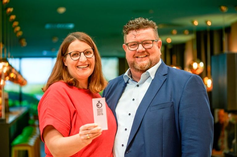 Two smiling people standing indoors; one is holding a small white card with a black apple symbol. The background references the German Design AWARD Special 2019 and features warm lighting and modern decor, radiating an elegant and contemporary atmosphere.