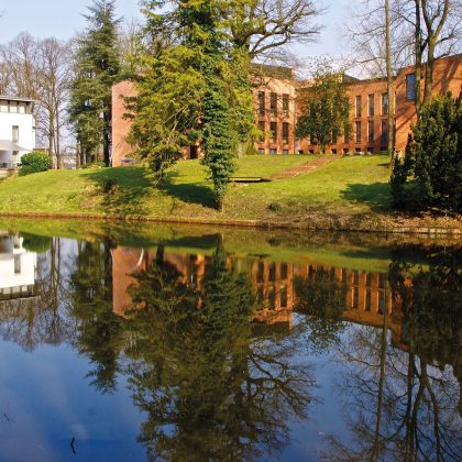Das moderne Backsteingebäude und die elegante weiße Struktur der Arcadeon Suite spiegeln sich wunderschön in einem Teich, umgeben von üppigen Bäumen und Gras unter einem klaren Himmel.