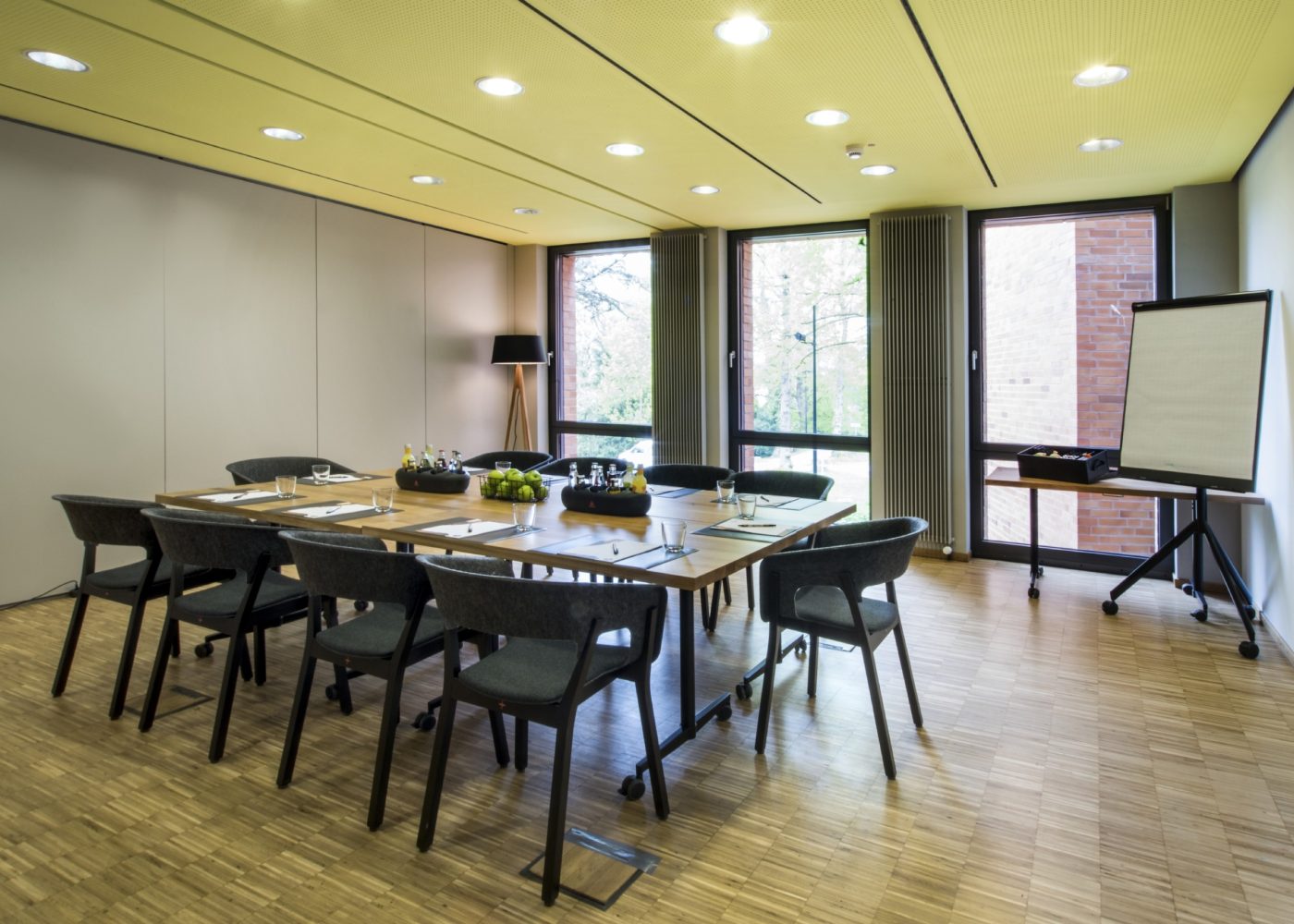 Ein moderner Konferenzraum verfügt über einen rechteckigen Tisch im Kombi-Stil für acht Personen, umgeben von eleganten Stühlen. Ein Flipchart steht bereit, während große Fenster einen ruhigen Blick auf Bäume bieten, die an die friedliche Atmosphäre in Cluny erinnern.