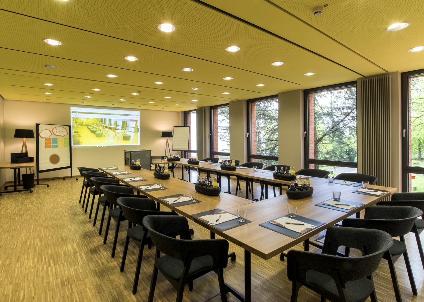 Ein Konferenzraum mit langen, U-förmig angeordneten Tischen, Stühlen und einer Projektionsleinwand, auf der eine Präsentation gezeigt wird, die an Clunys Erbe erinnert. In der Nähe steht ein Flipchart und große Fenster bieten einen ruhigen Blick auf die Bäume draußen.