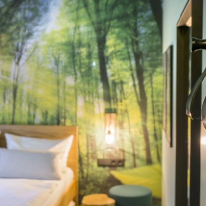 Eine Schlafzimmereinrichtung mit einem Wandbild zum Thema Wald, einem ordentlich gemachten Bett, einer Wandleuchte an der Decke und einem dekorativen Wandhaken von ARCADEON im Vordergrund.