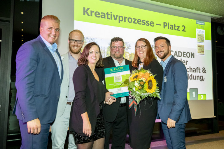 A group of six people poses smiling and holding flowers in front of a screen displaying "Kreativprozesse – Platz 2" at ARCADEON. This celebration follows the announcement of their achievement at one of the most prestigious conference hotels in Germany.